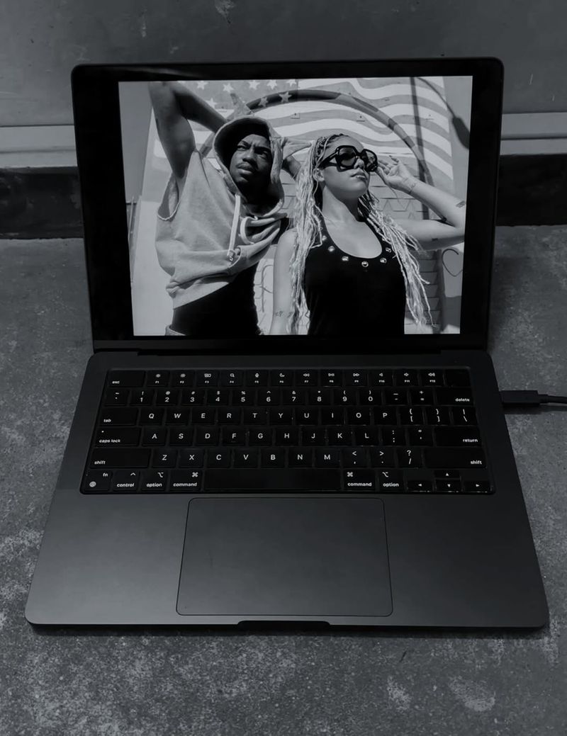 Monochrome shot of a laptop displaying a man and woman, with an American flag backdrop, against a plain wall and floor.