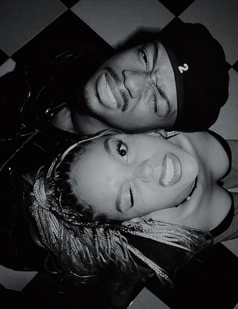Monochromatic close-up of a smiling adult man and woman with braided hair. Both are facing the camera against a checkerboard background.
