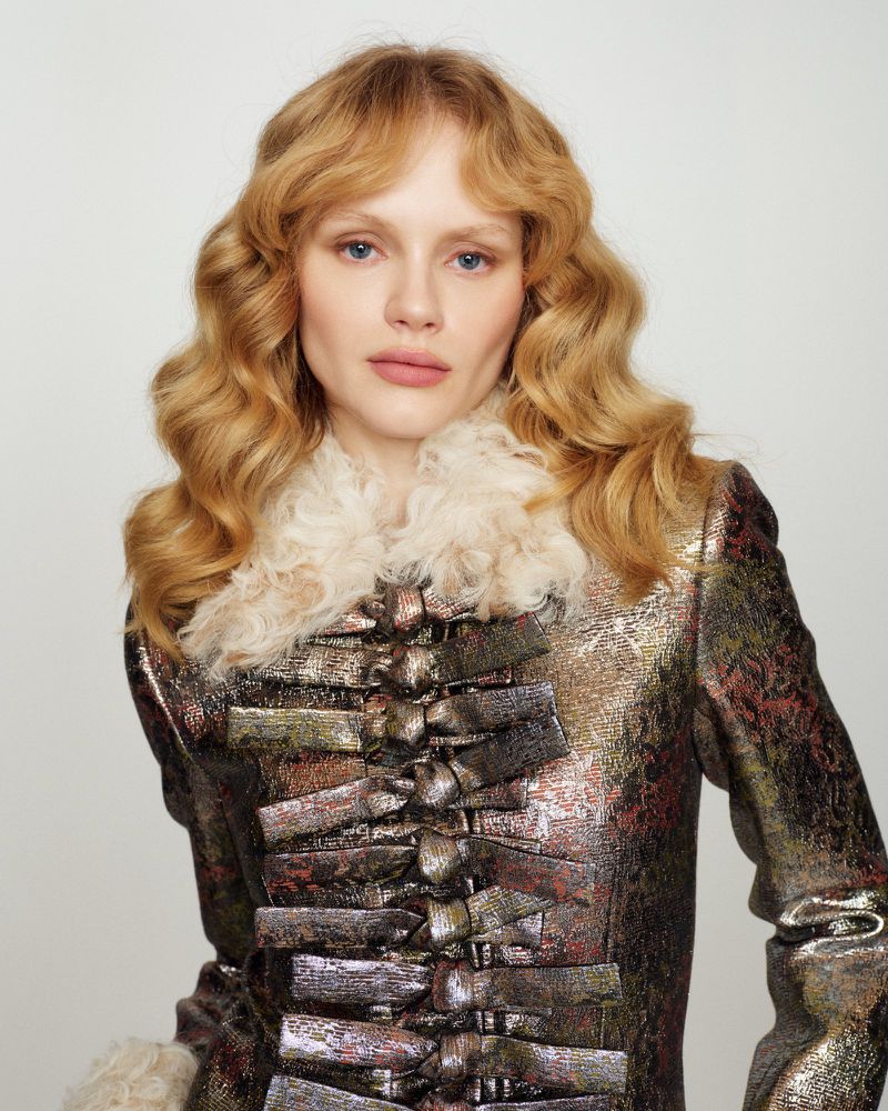 Emma Laird wearing a metallic patterned jacket with a cream shearling collar in an editorial studio portrait.