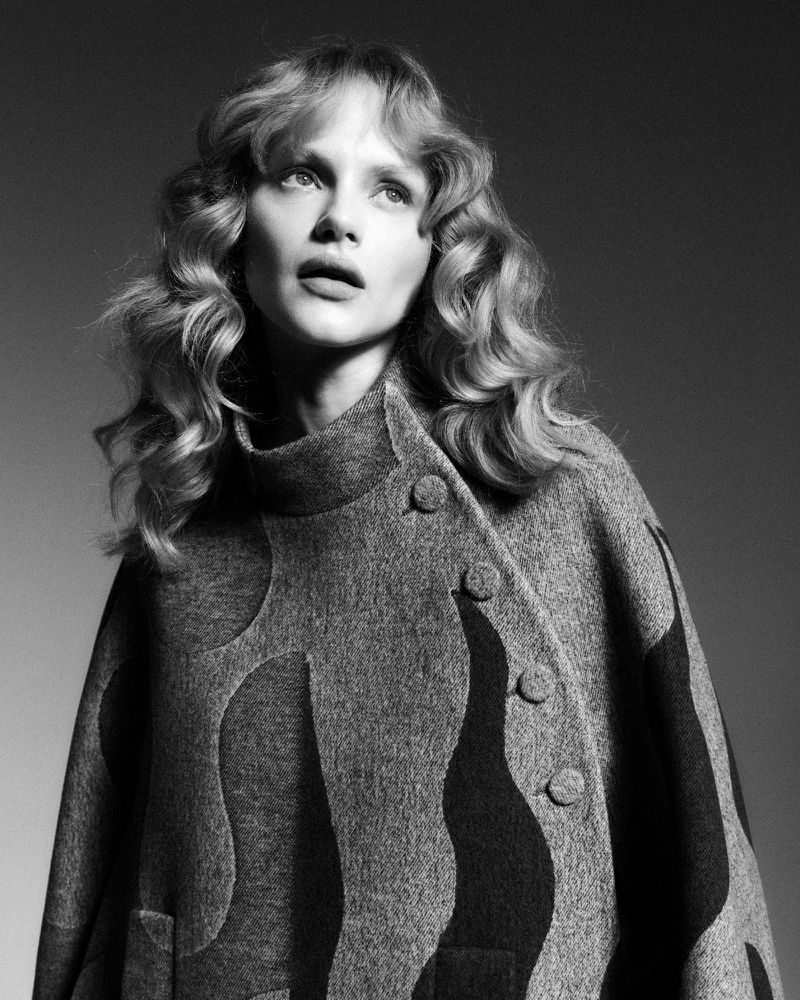 Black and white portrait of a blonde woman wearing a wool blend textured pattern coat, gazing upward in a neutral gray background.