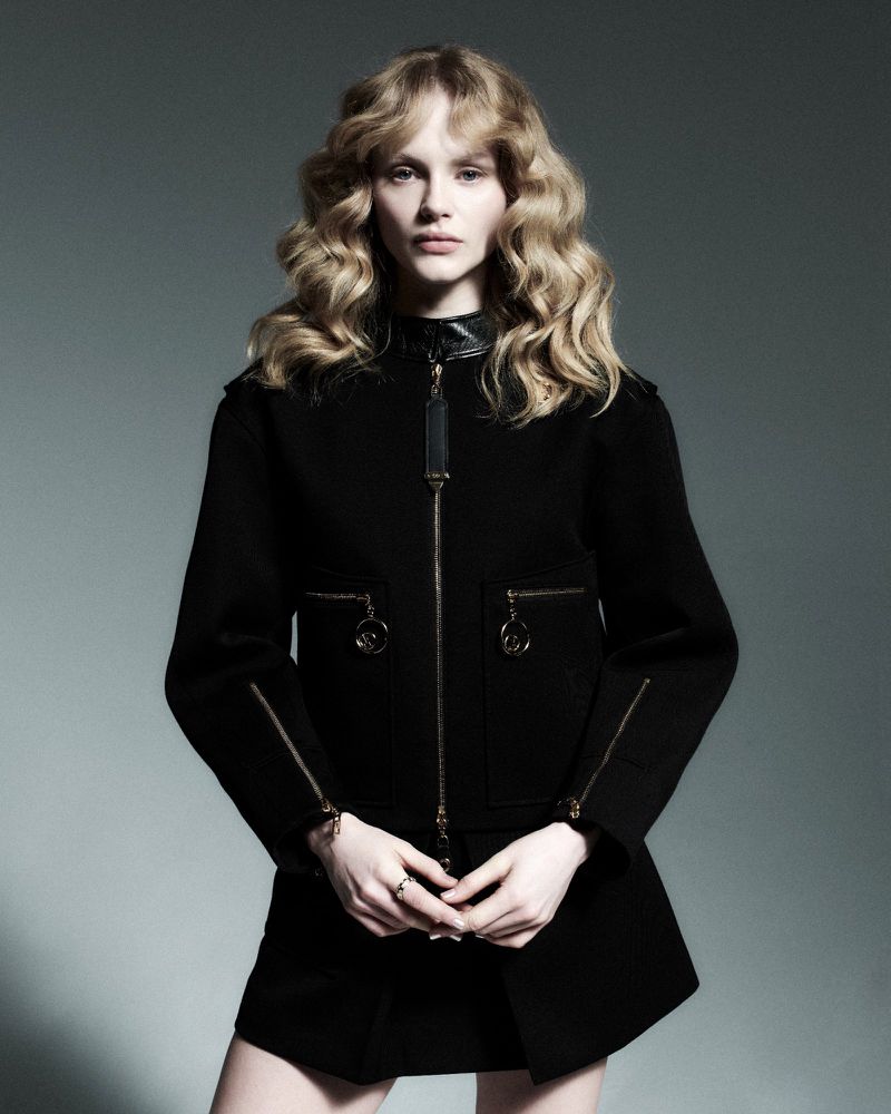 Fashionable woman with curly hair wearing a black jacket and skirt posing in a photo studio against a gray background.