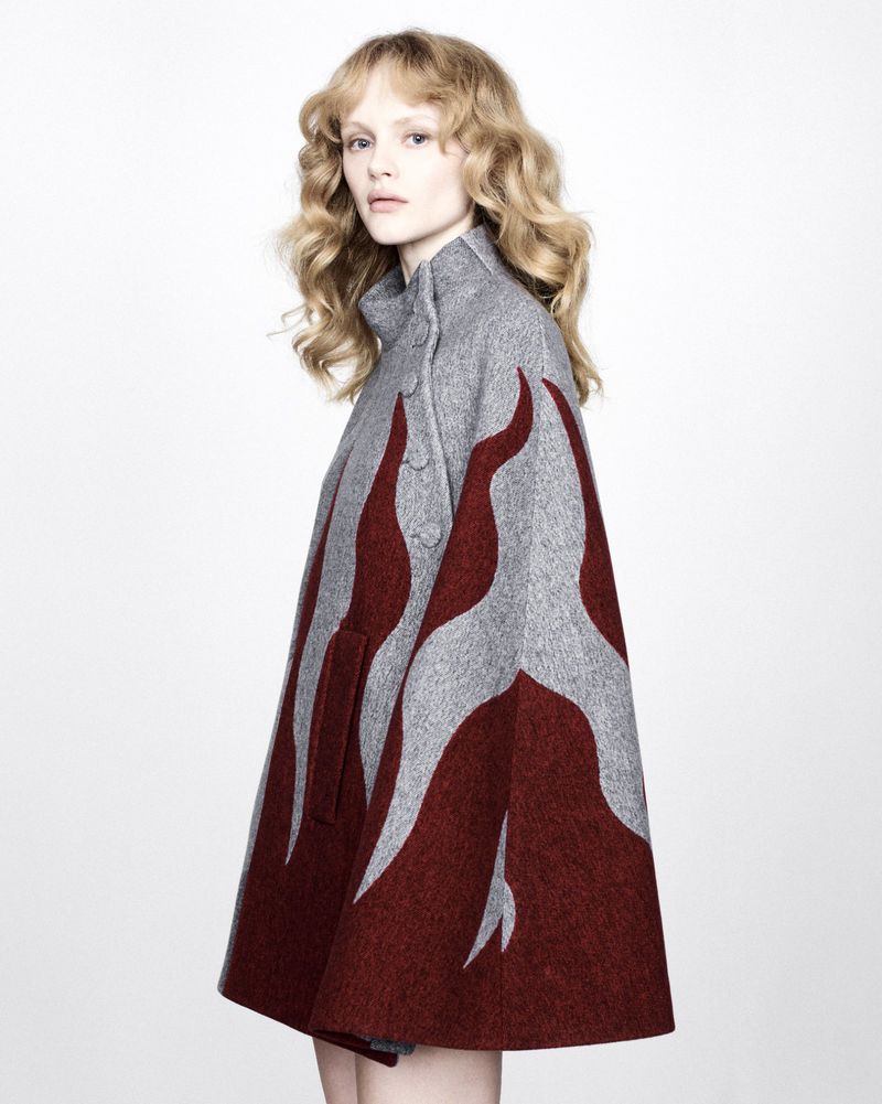 Woman wearing a gray cape-style coat with abstract red flame design, posing in a studio with soft lighting and light hair.