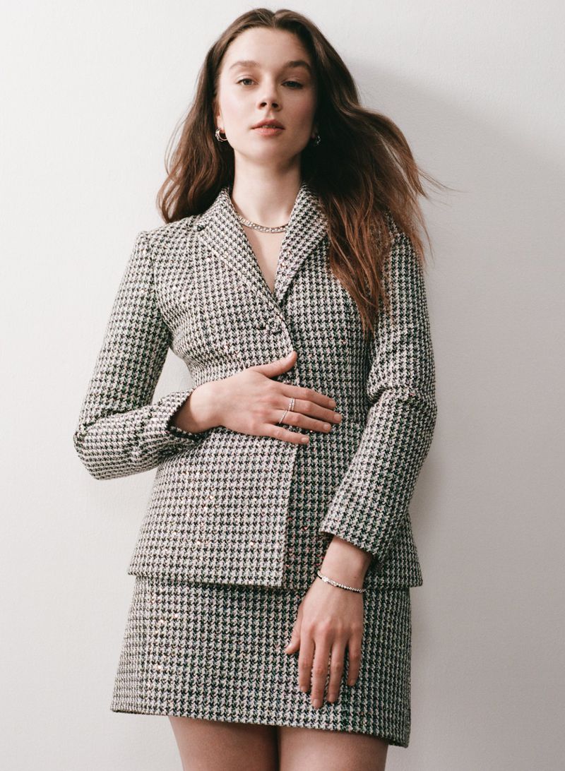 Young woman in a grey houndstooth blazer and skirt poses indoors against a white wall, looking directly at the camera. Fashion shoot.
