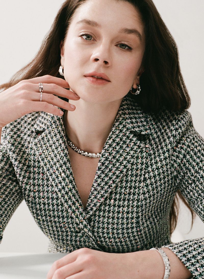 Adult female wearing a houndstooth blazer with silver jewelry, including a necklace, earrings, rings, and a bracelet, looking at the camera.
