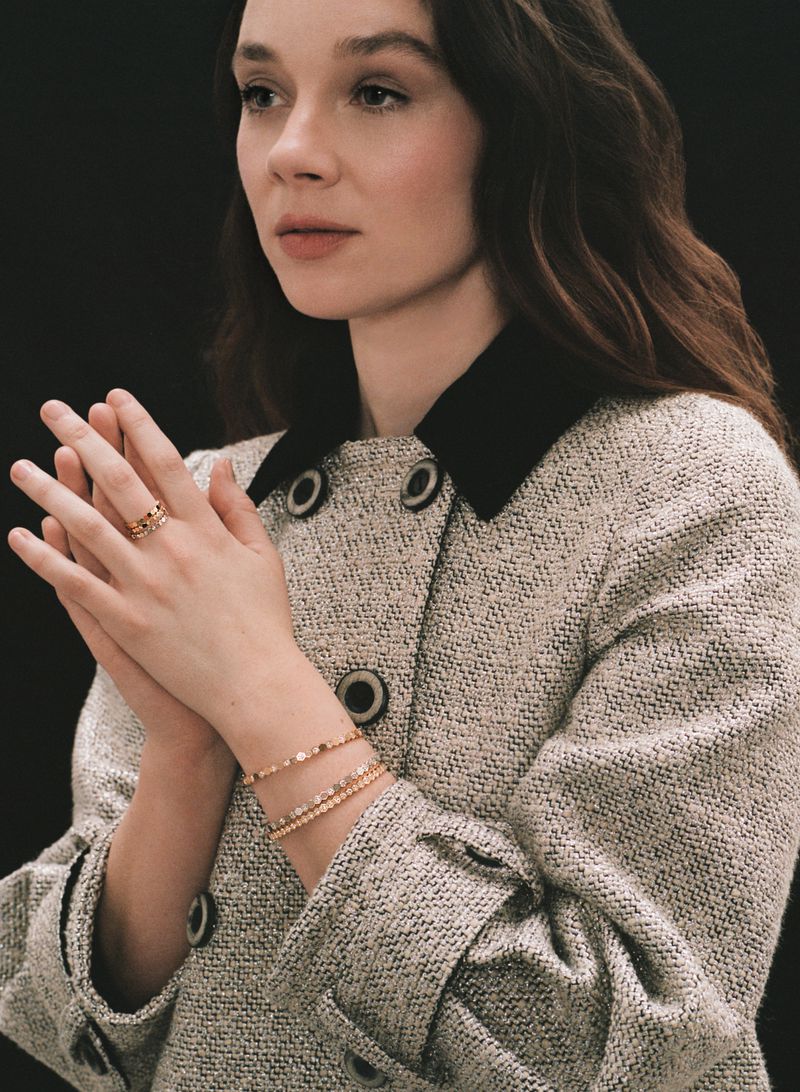 Portrait of an adult woman with brown hair wearing a tweed jacket, gold bracelets, and a ring. She has her hands together.