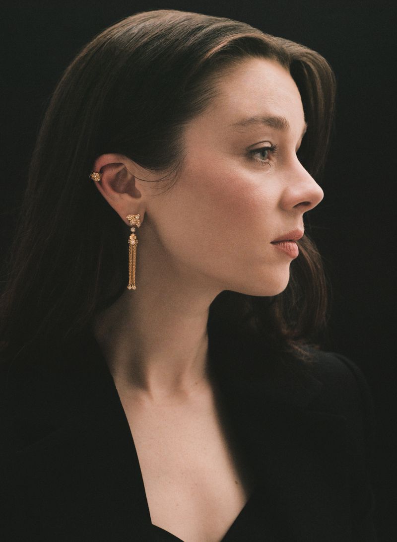 Profile portrait of a young adult woman with long brown hair wearing gold earrings and a black jacket against a dark background.