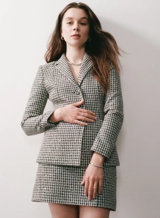 Claudia Jessie wearing a grey houndstooth blazer and matching mini skirt against a white studio background.