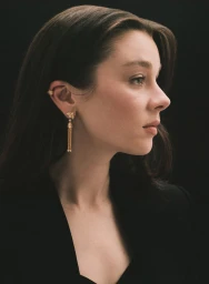 Side profile portrait of actress Claudia Jessie wearing gold tassel drop earrings against a solid black background.