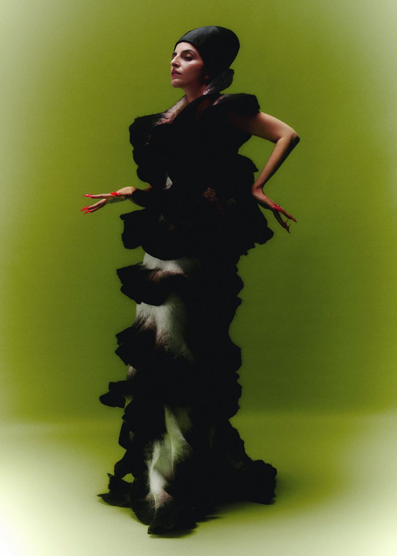 A fashionable woman in an elaborate ruffled black gown and head covering poses gracefully against a soft green backdrop in a studio setting.