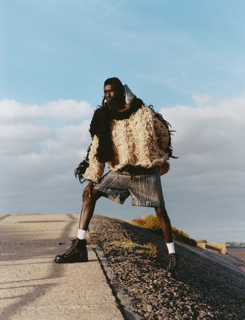 Model Jum Kuochnin standing in an oversized textured knit sweater, shorts, and black boots on an embankment against a blue sky.