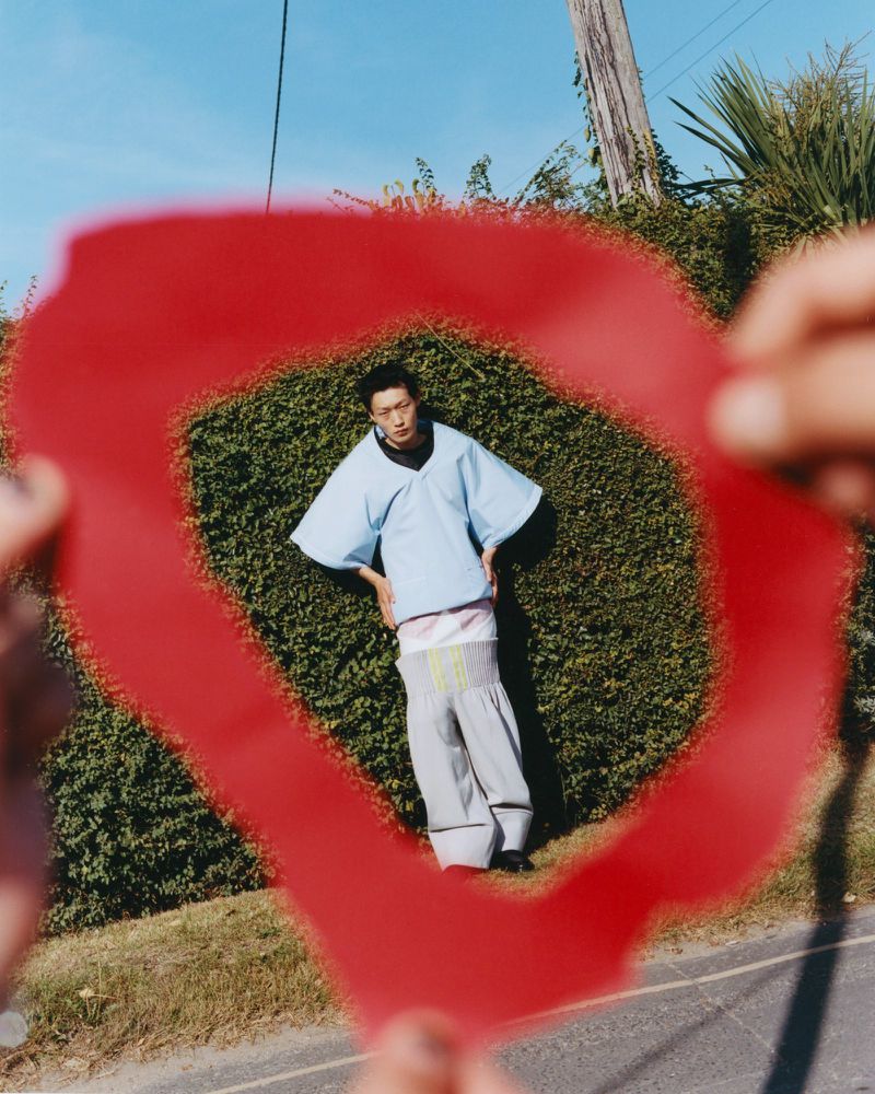 Fashion shot of an Asian adult male posing, framed by hands holding a red plastic sheet, against a background of lush green foliage.