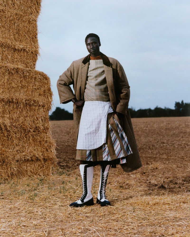 Dark-skinned adult poses in a field wearing a brown coat, layered skirts, knee-high boots, and standing next to a stack of hay bales.