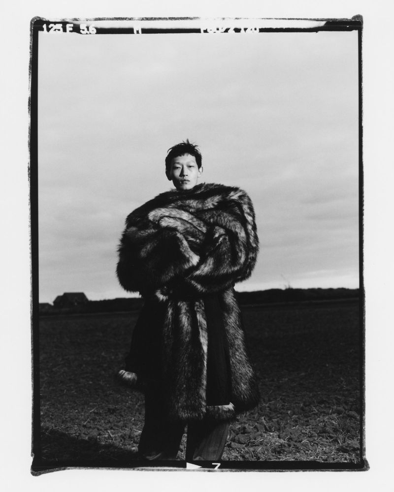 Monochrome portrait of a person in a fur coat standing in a field under a cloudy sky, creating a striking and high-contrast image.