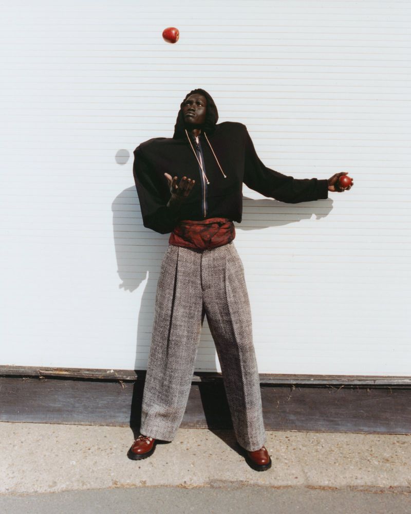 Adult male juggles apples in an outdoor setting, wearing a black hooded jacket, oversized grey pants, and dark brown dress shoes.