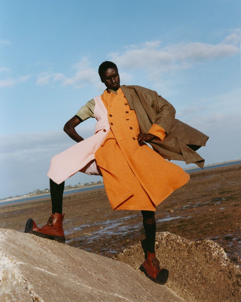 A fashion shot features a Black model in a multi-colored coat and brown boots, standing on a rocky landscape against a blue sky.