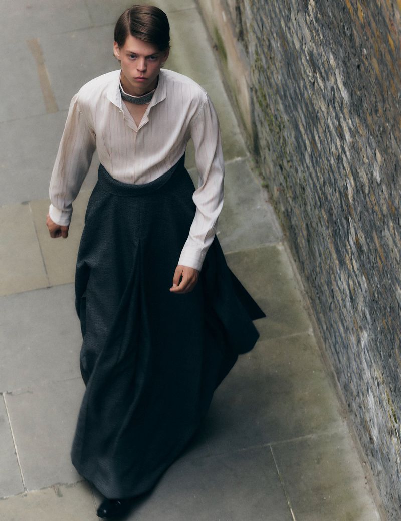 A solitary person in a white shirt and long, full dark skirt stands on a stone path alongside a textured stone wall in natural light.