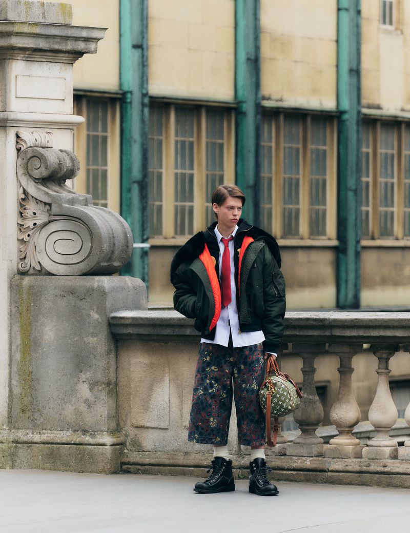 A young man stands on a balcony in a green bomber jacket, floral pants, and combat boots, holding a patterned satchel bag.