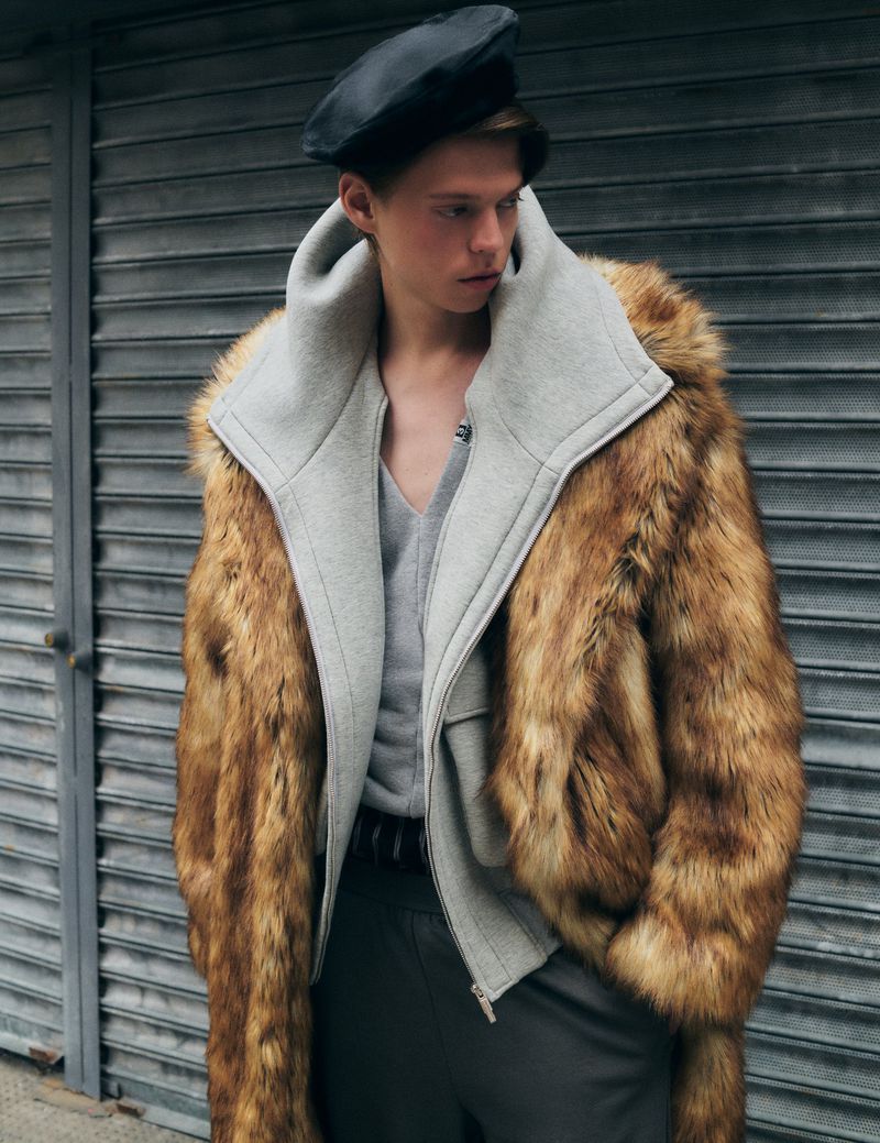 Male fashion model wears a fur coat with grey lining and a black beret against a metal background in a medium shot.