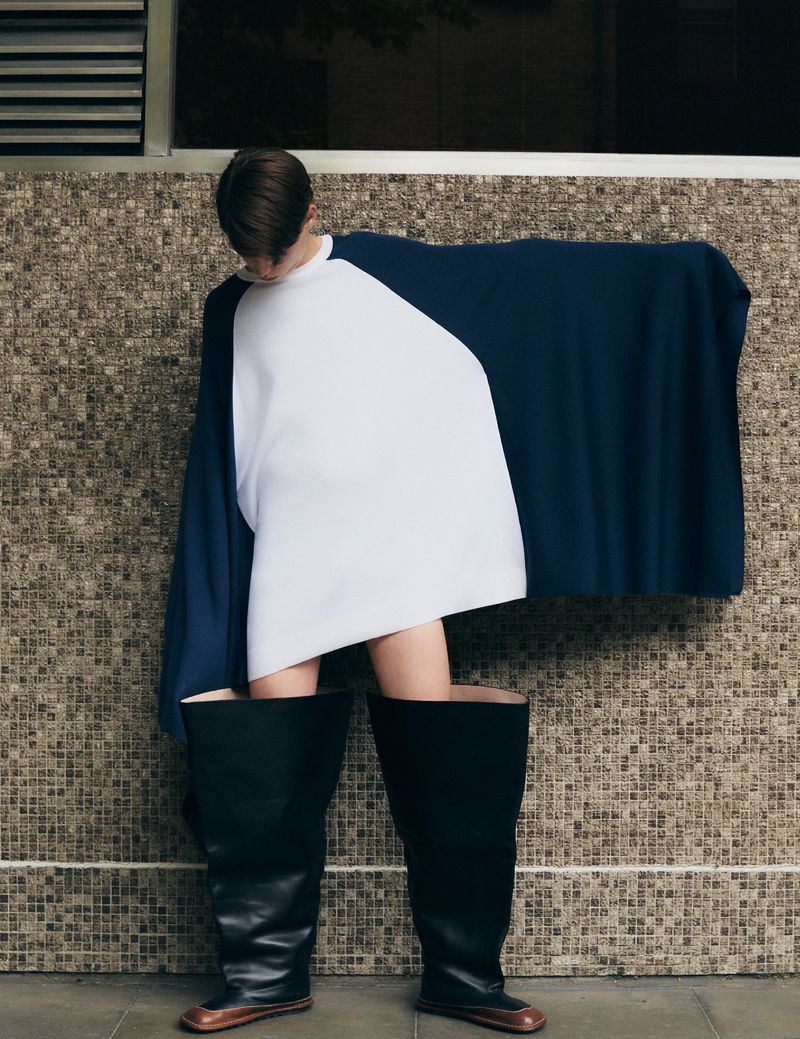 Fashion shot of a person wearing oversized blue and white top, standing in large black boots, posing against a textured wall indoors.