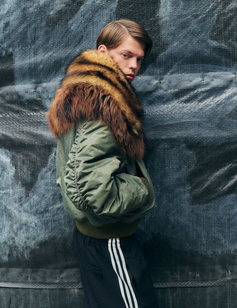 Fashion shot of a young adult male wearing a fur collar bomber jacket and athletic pants standing against a textured gray wall.