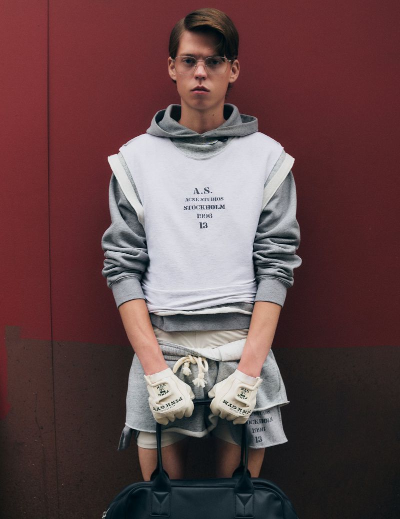 Male model poses against a two-toned red and brown background in a gray hoodie, white top, gray shorts, gloves, and black duffel bag.