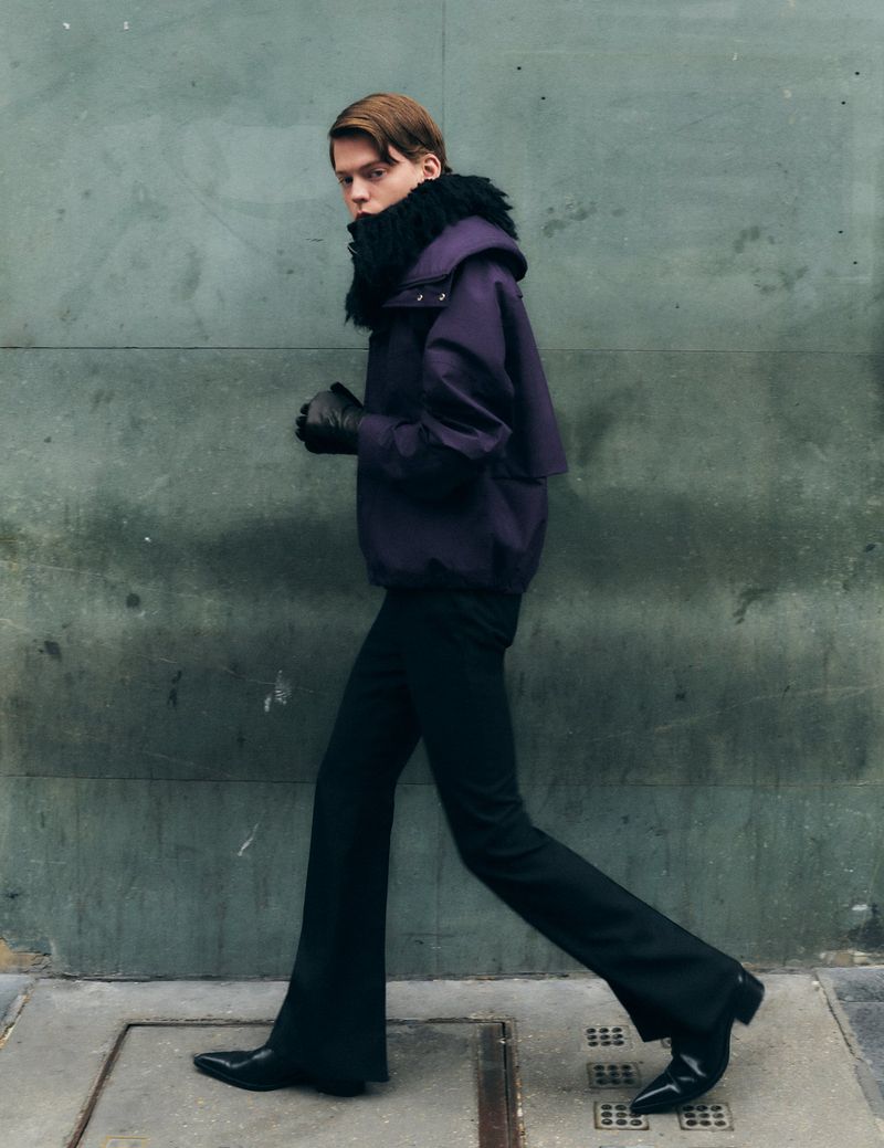 A young adult man with dark hair walks along a city sidewalk wearing a purple winter coat, black pants, and black boots.