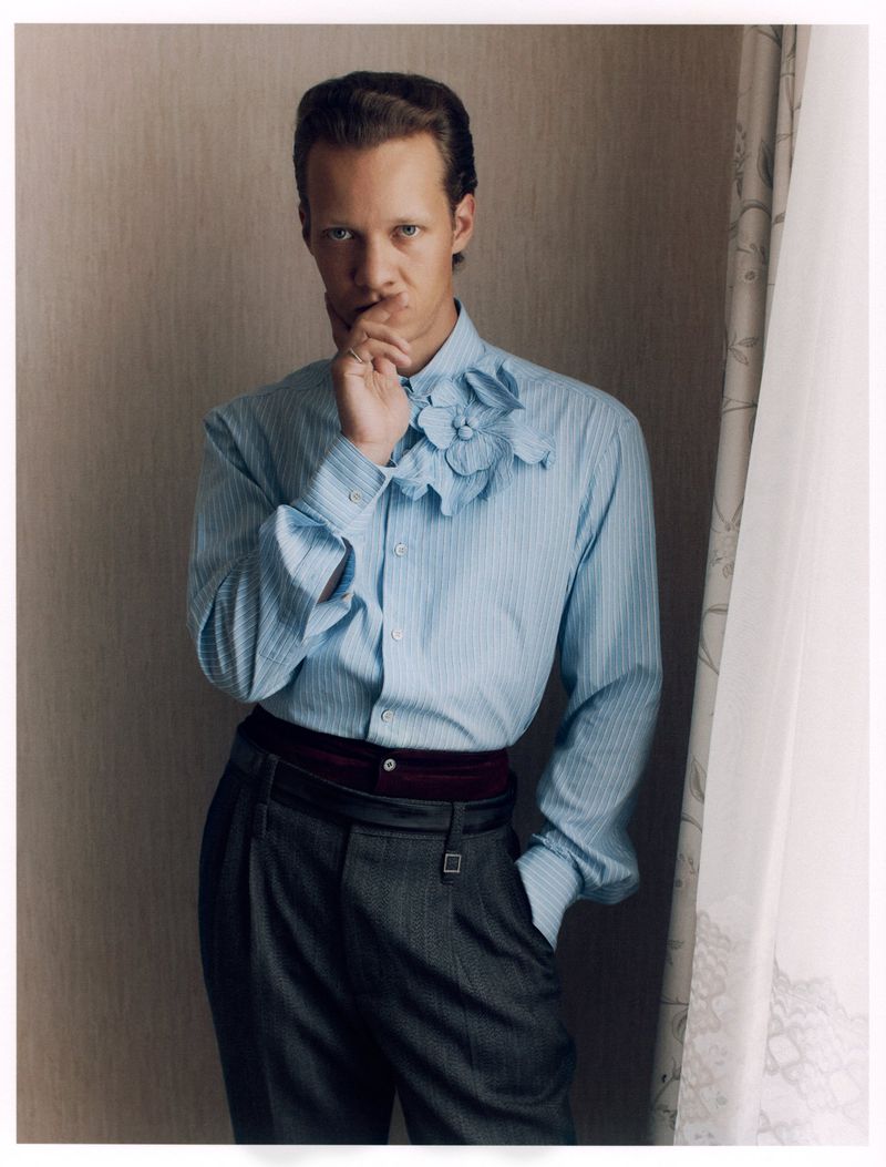 Fashionable adult male poses in striped shirt and pleated pants against neutral backdrop with soft window light, in a studio setting.