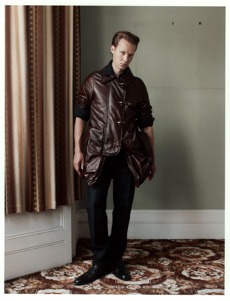Full-length shot of a young adult man in a brown leather jacket, black pants, and black shoes, standing indoors with patterned carpet and a curtain.