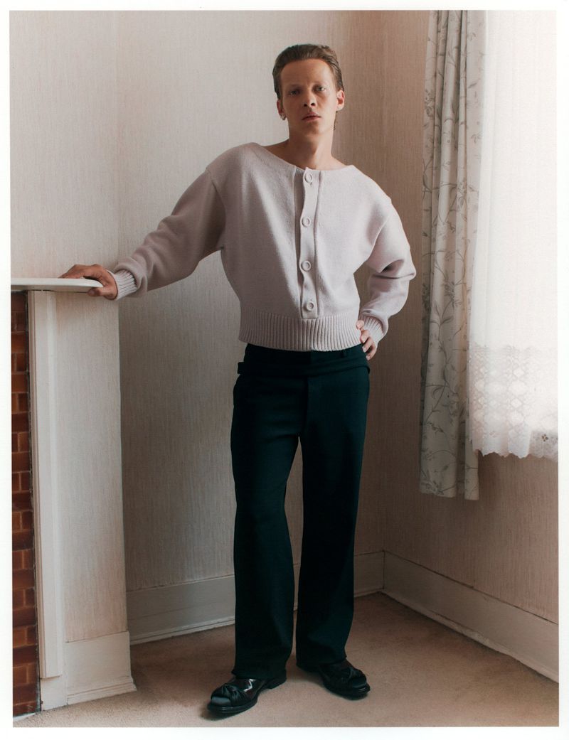Full-length portrait of a young fair-skinned male model wearing a cardigan and slacks, posing indoors against a neutral wall.