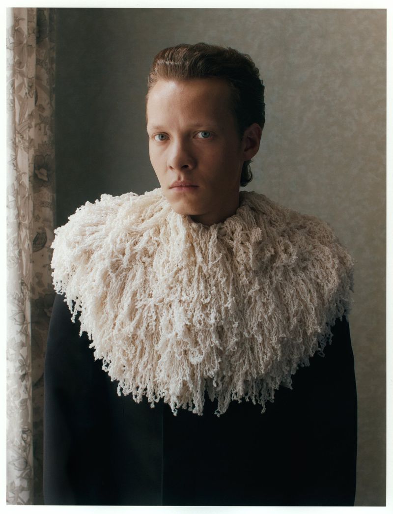 Portrait of a person with fair skin and slicked-back brown hair wearing a black jacket and a white yarn ruff collar in an indoor setting.