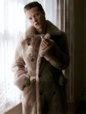 Actor Felix Kammerer posing in a plush beige shearling double-breasted coat beside a sheer curtained window.