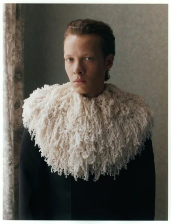 Actor Felix Kammerer wearing an oversized textured cream knit collar and black jacket against a soft interior background.