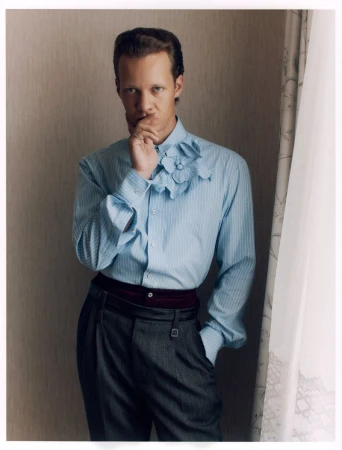 Felix Kammerer posing in a light blue floral-embellished shirt and dark pleated trousers by a curtained window.