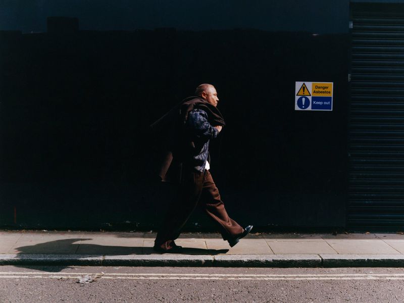 An adult man walks on a city sidewalk next to a dark wall with a caution sign, wearing brown pants, a patterned shirt, and carrying a coat.