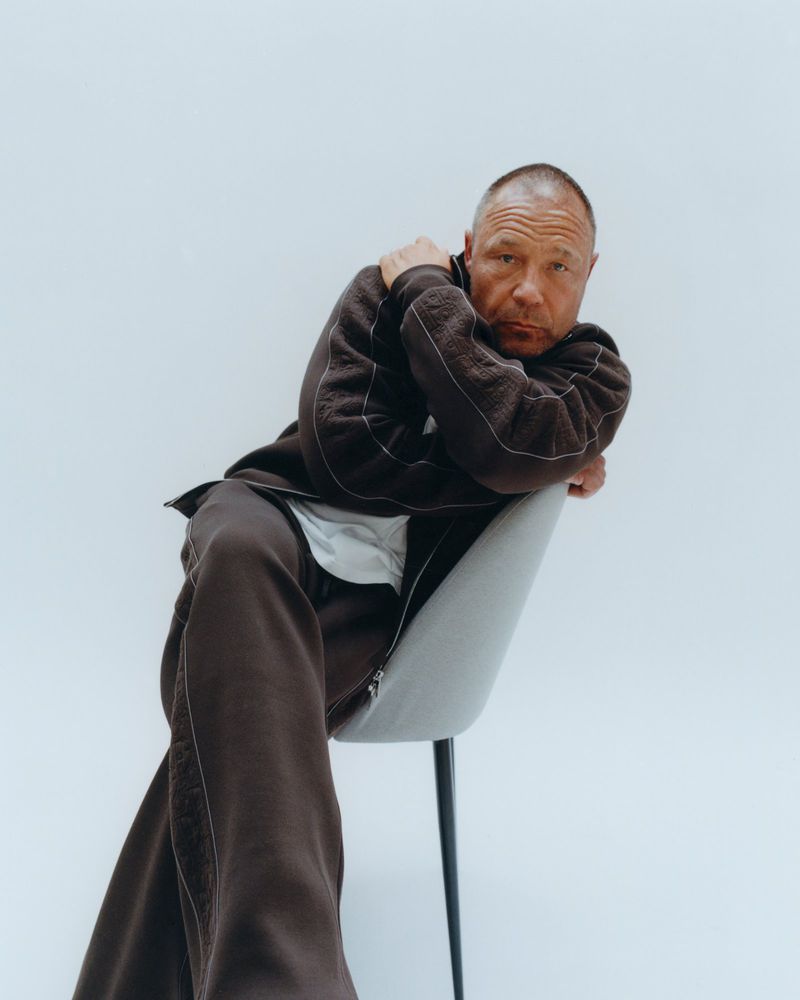 Portrait of a mature man seated on a gray chair in a photo studio, wearing a brown tracksuit and leaning forward, with a direct gaze.