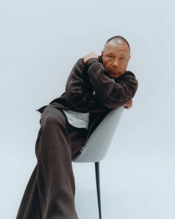 Portrait of a mature man seated on a gray chair in a photo studio, wearing a brown tracksuit and leaning forward, with a direct gaze.