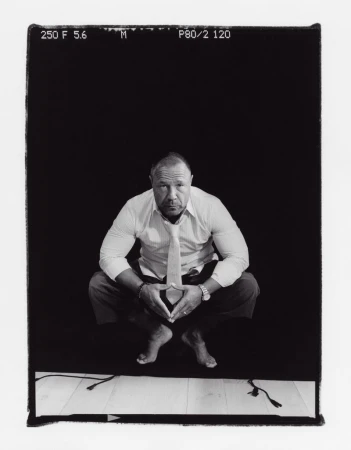 Monochrome portrait of a man in a light-colored shirt and tie, squatting barefoot against a black background, his hands touching.