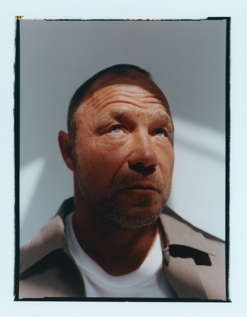 Close-up portrait of actor Stephen Graham looking upwards in direct sunlight wearing a white shirt and taupe overshirt.