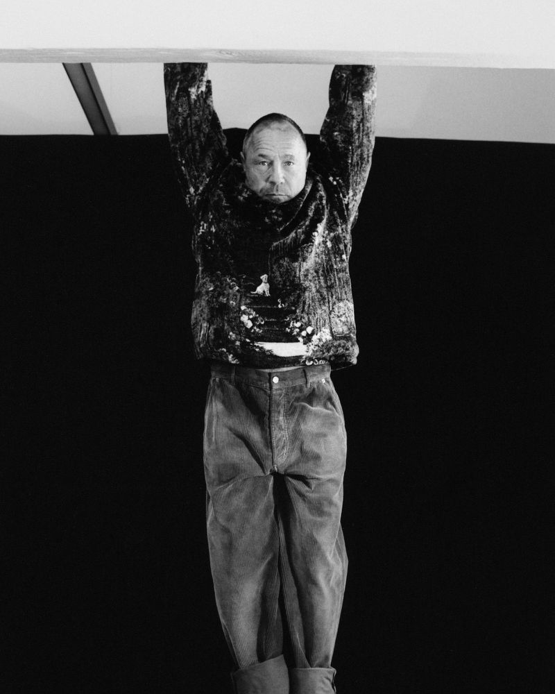 Black and white studio portrait of a man hanging from an overhead bar, wearing a patterned shirt and corduroy pants, against a dark backdrop.