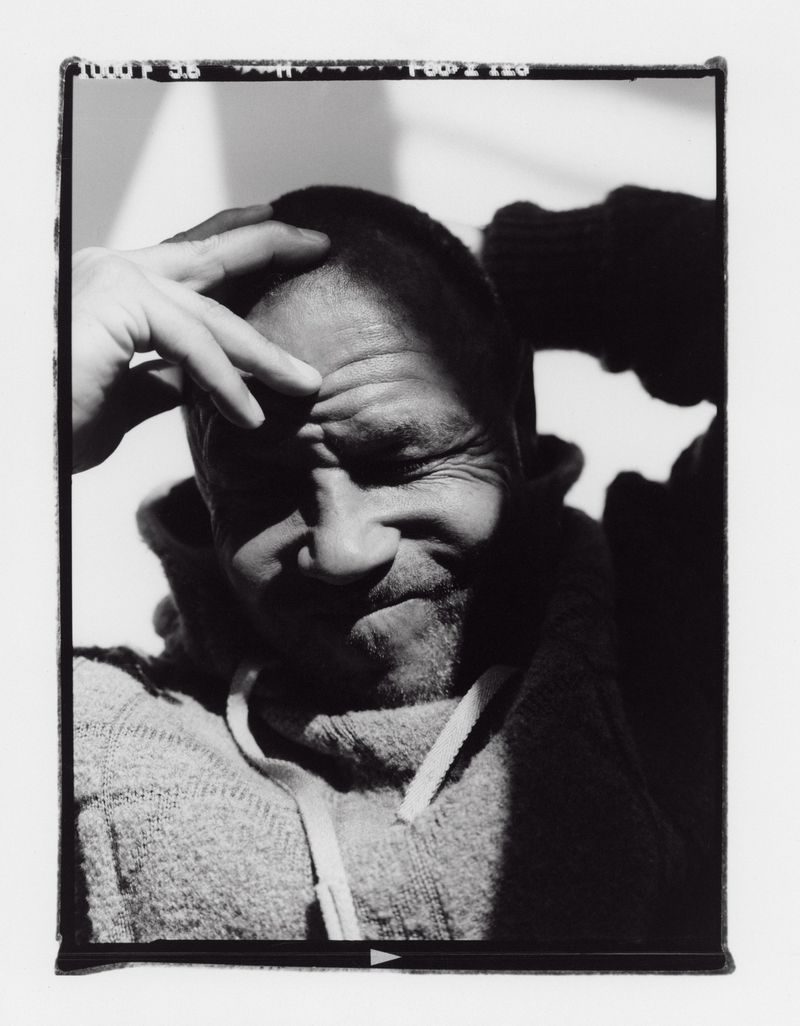 Black and white portrait of adult male in a hooded sweatshirt with hand on head, partially shadowed against a light background.