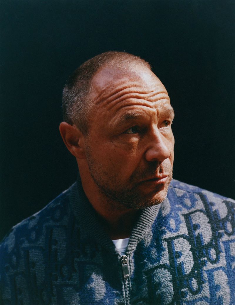 Portrait of a mature adult male with a bald head and stubble beard, wearing a patterned blue zip-up jacket against a dark background.