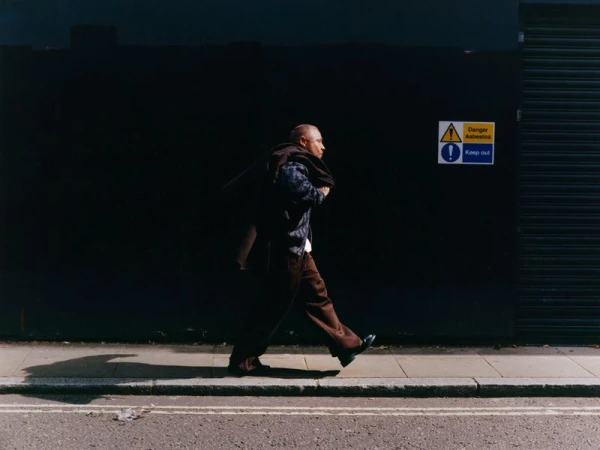 Actor Stephen Graham walking along a sidewalk in front of a dark wall with a brown jacket over his shoulder.