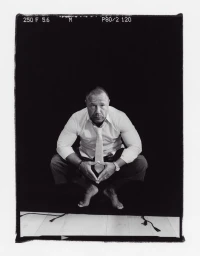 Stephen Graham crouching barefoot in a collared shirt, necktie, and trousers against a dark backdrop in a film frame.