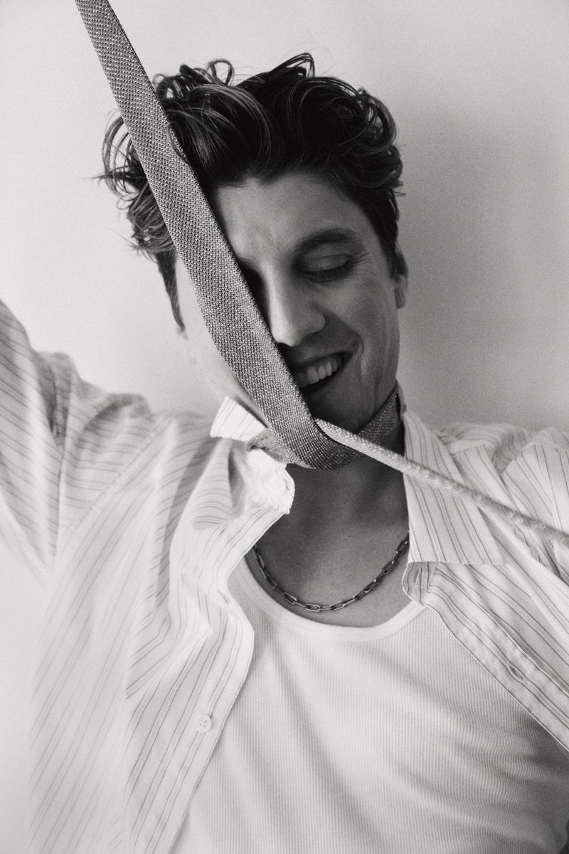 Portrait of actor Lukas Gage smiling with a strap across his face wearing an open striped shirt and chain necklace against a light background