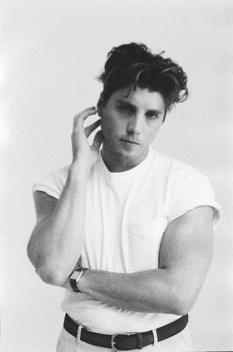 Black and white portrait of a young adult male wearing a t-shirt, his hand near his face, captured in a medium shot against a plain background.