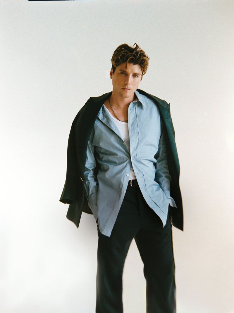 Portrait of a blonde male model wearing a light blue unbuttoned shirt with a dark green blazer draped over his shoulders in a studio.