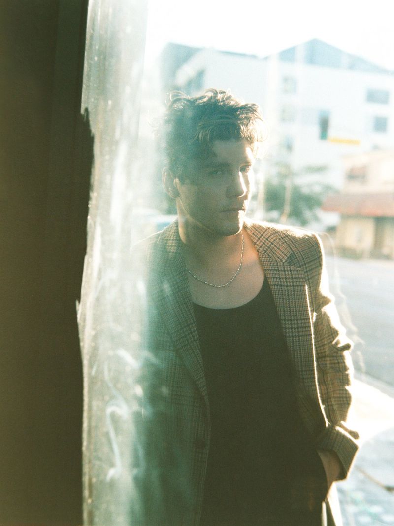 Actor Lukas Gage in a plaid blazer and black tank top standing behind a reflective glass window in natural light.