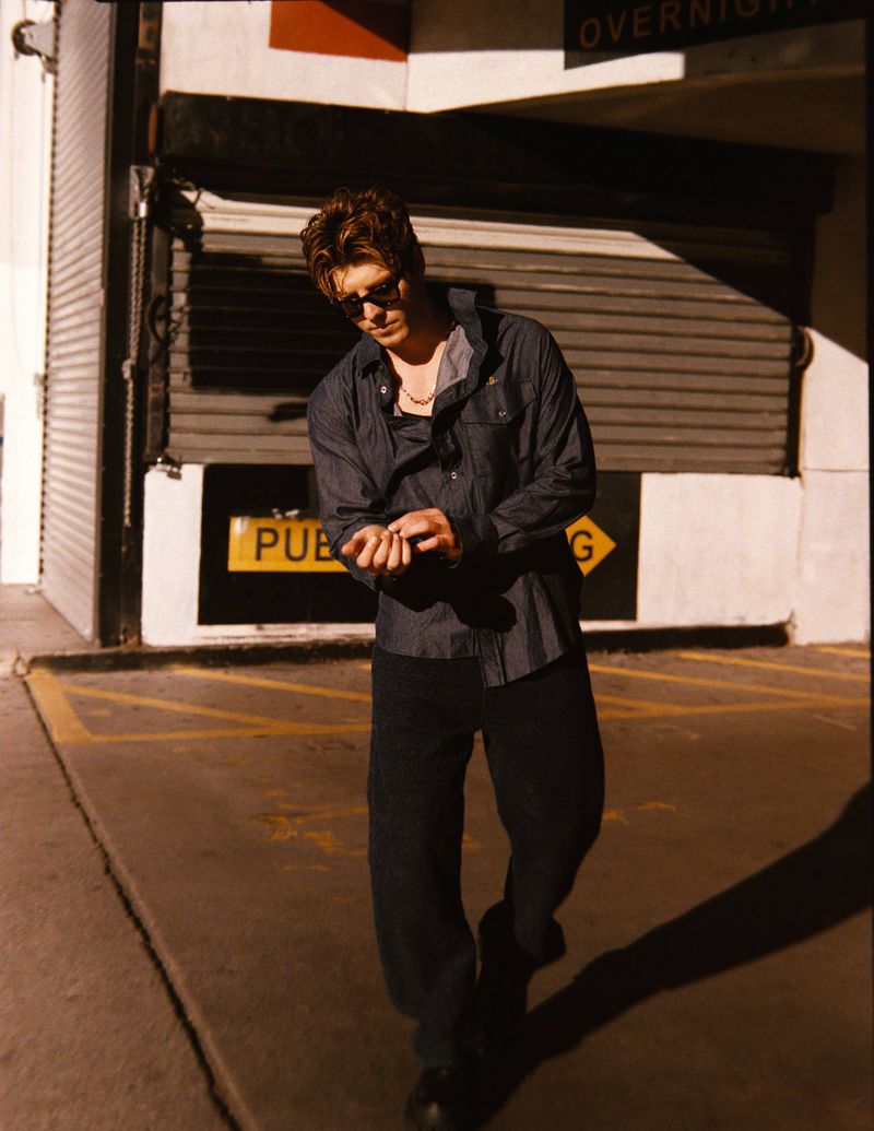 Man in sunglasses and unbuttoned shirt stands in an urban alleyway with a garage door. Moody lighting, casual fashion.
