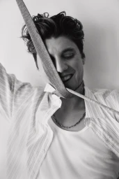 Portrait of actor Lukas Gage smiling with a strap across his face wearing an open striped shirt and chain necklace against a light background