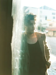 Actor Lukas Gage in a plaid blazer and black tank top standing behind a reflective glass window in natural light.